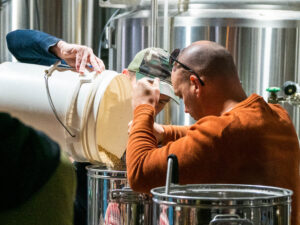 International Homebrew Day - Masterclass- Man in Orange shirt pouring a bucket in to a homebrew kit
